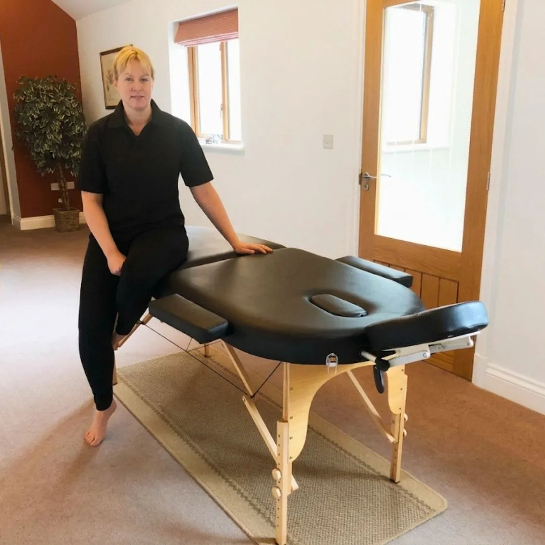 Porta-Lite Oval portable massage table featuring German beech wood legs, a 5cm double-layer high-density foam top, and a 500lbs cable-lock load capacity; shown in a home treatment setup with removable side armrests and a front arm shelf to solve the problem of restricted movement and bruised shins in small rooms by letting therapists in Nottingham flow smoothly around the corner-free layout.