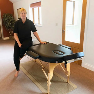 Porta-Lite Oval portable massage table featuring German beech wood legs, a 5cm double-layer high-density foam top, and a 500lbs cable-lock load capacity; shown in a home treatment setup with removable side armrests and a front arm shelf to solve the problem of restricted movement and bruised shins in small rooms by letting therapists in Nottingham flow smoothly around the corner-free layout.
