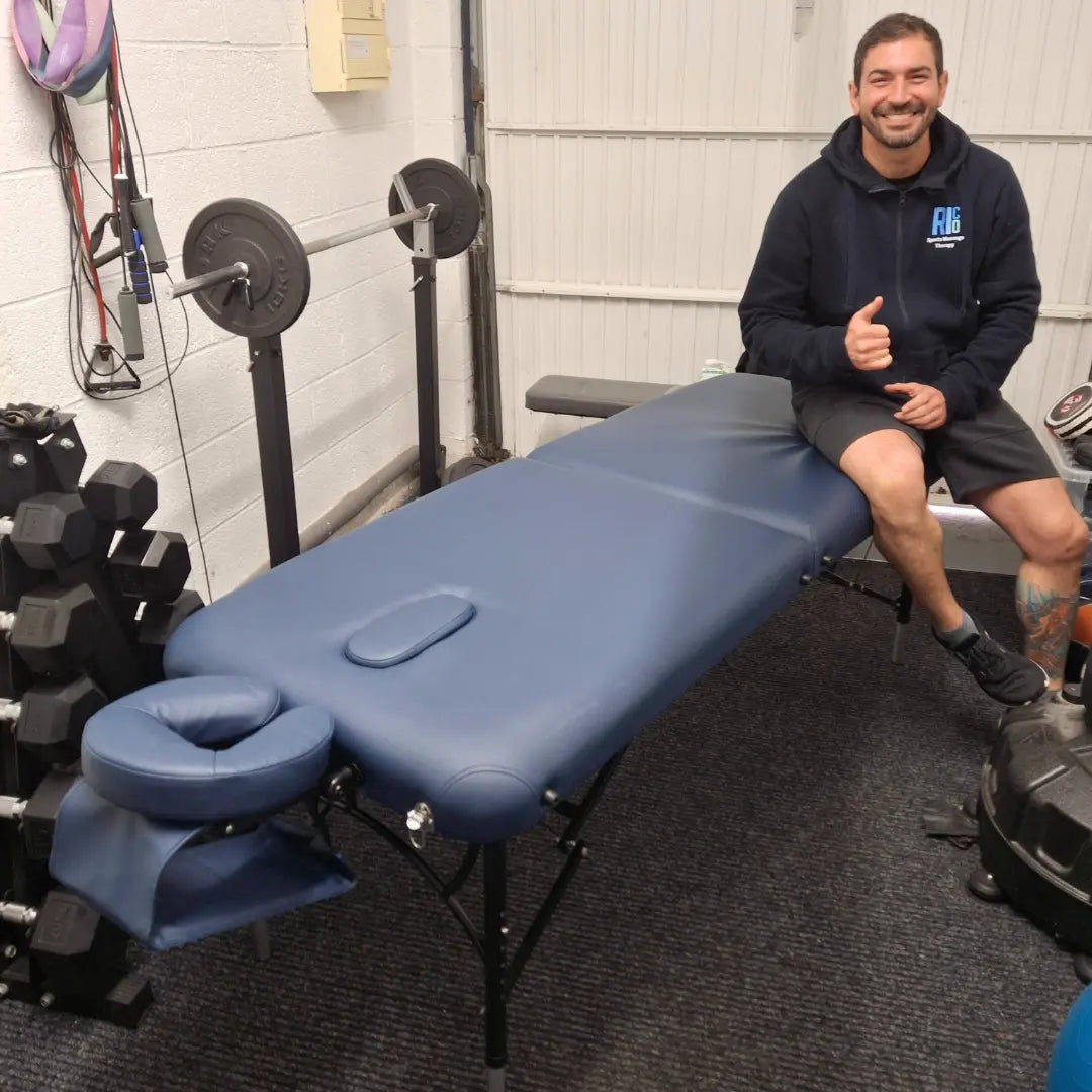 A professional therapist giving a thumbs up while sitting on a navy blue BodyPro Active portable massage table. The table features a lightweight 12kg aluminum frame, 7cm high-density multi-layer foam, and a professional headrest with an arm sling, shown in a Sheffield gym setting for sports therapy. Available in the United Kingdom and Ireland.