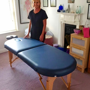 Porta-Lite Oval portable massage table in navy blue, constructed with sturdy German beech wood legs, a stable cable tension chassis, and an integrated face hole; shown in an at-home treatment room in Cardiff to solve the problem of navigating tight domestic spaces by eliminating sharp, square corners that catch clothing, bruise knees, and restrict smooth therapist movement during high-volume daily treatments.
